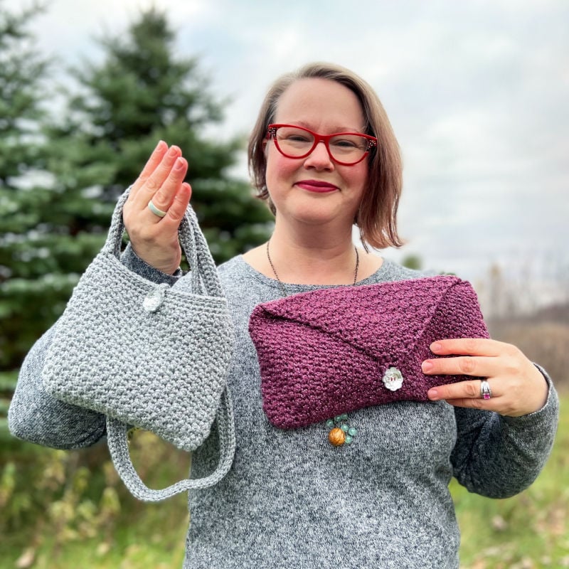 Designer holding up a crocheted silver shoulder bag and a crocheted purple clutch purse