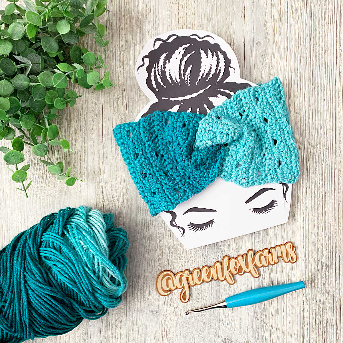 crochet ear warmer free pattern flat lay image of teal yarn, ear warmer on head cutout, with leaves and crochet hook